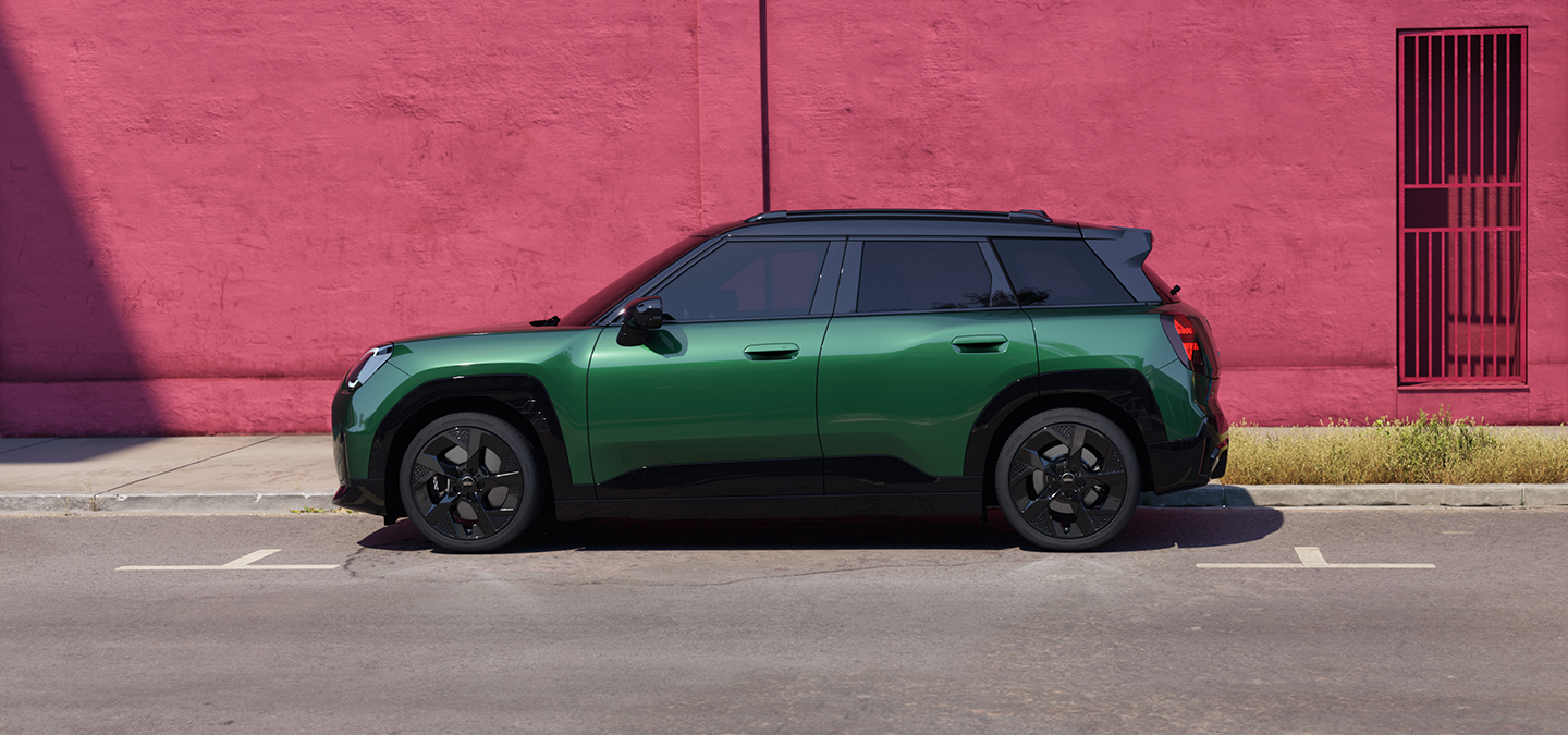 Sānu skats uz pilnībā elektrisko MINI Aceman British Racing Green krāsā ar melnu jumtu, kas novietots uz ielas.