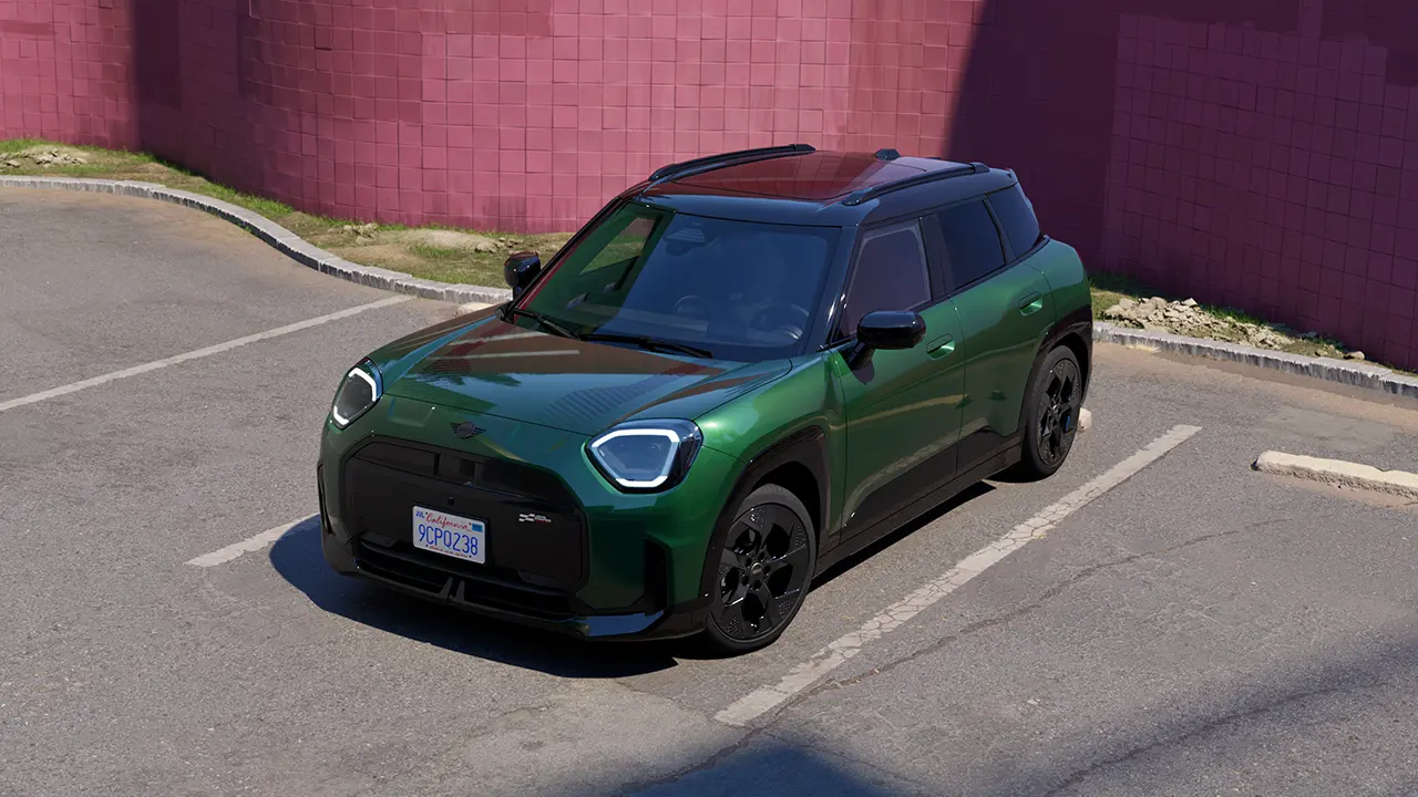 Priekšpuses skats – stāvvietā novietots pilnībā elektriskais MINI Aceman British Racing Green krāsā, fonā rozā siena