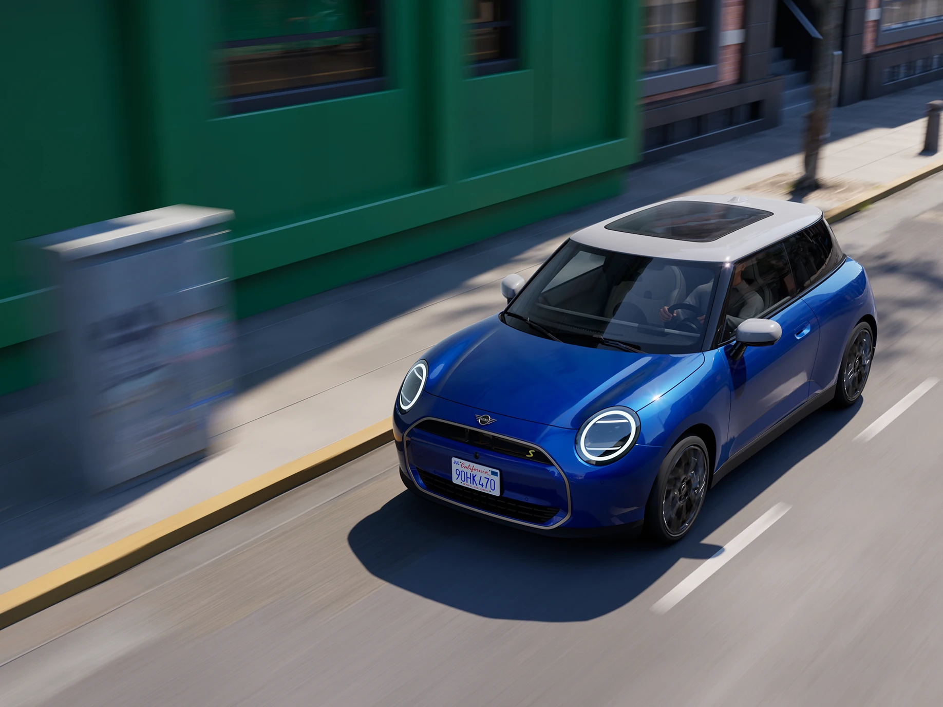 Skats no augšas uz pilnībā elektrisko MINI Cooper Blazing Blue krāsā ar baltu jumtu, braucot pa ielu.