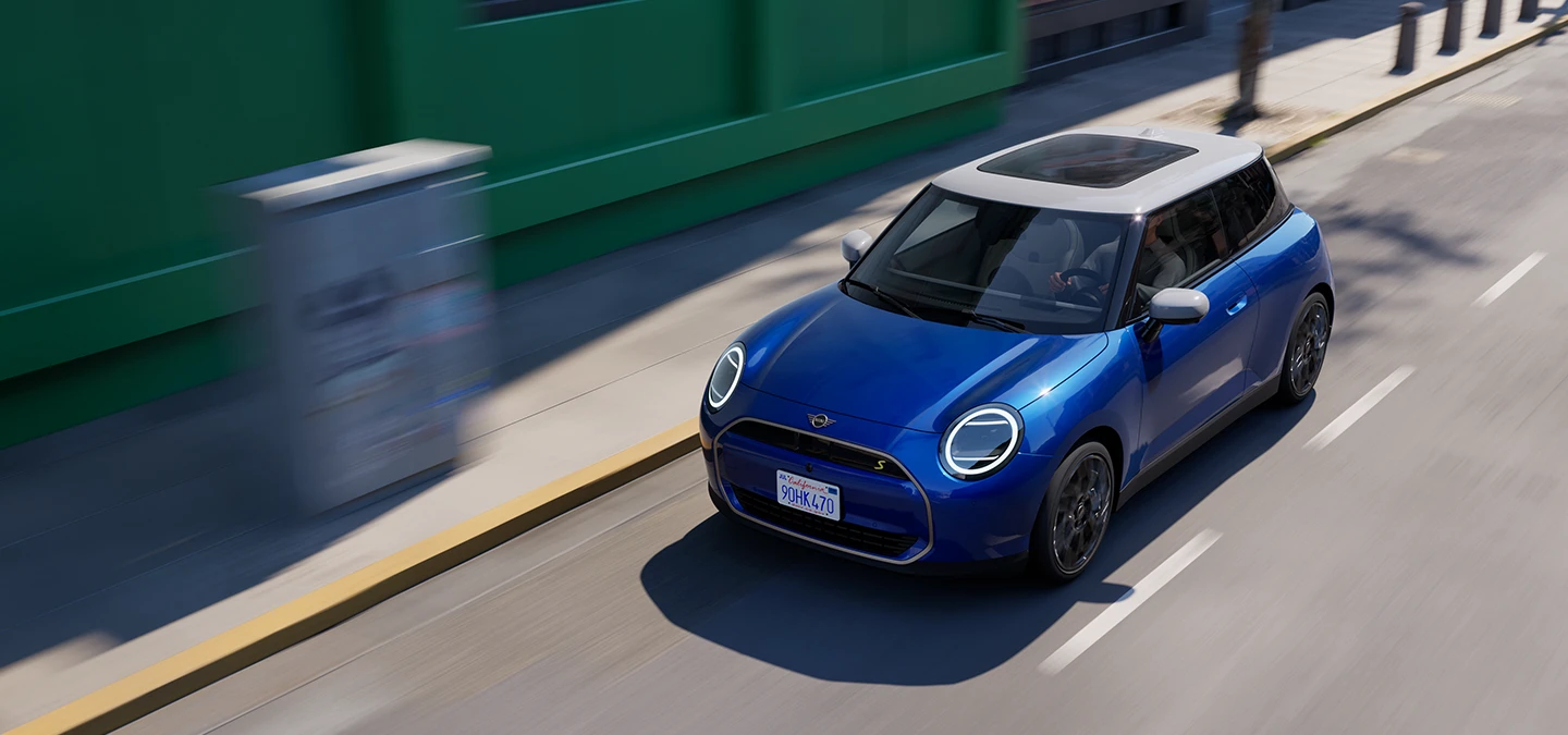 Skats no augšas uz pilnībā elektrisko MINI Cooper Blazing Blue krāsā ar baltu jumtu, braucot pa ielu.