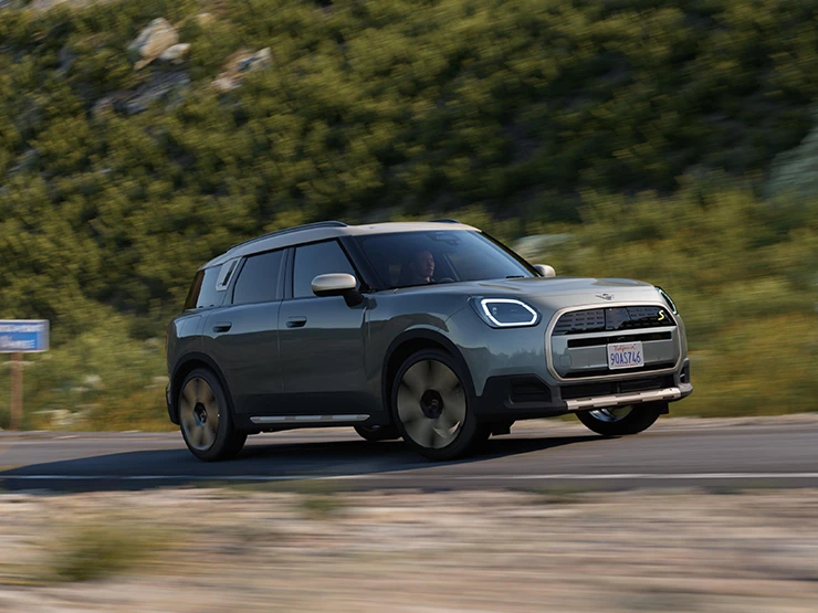Sānskats uz pilnībā elektrisko MINI Countryman Smokey Green krāsā ar Vibrant Silver jumtu, braucot bezceļa apstākļos.