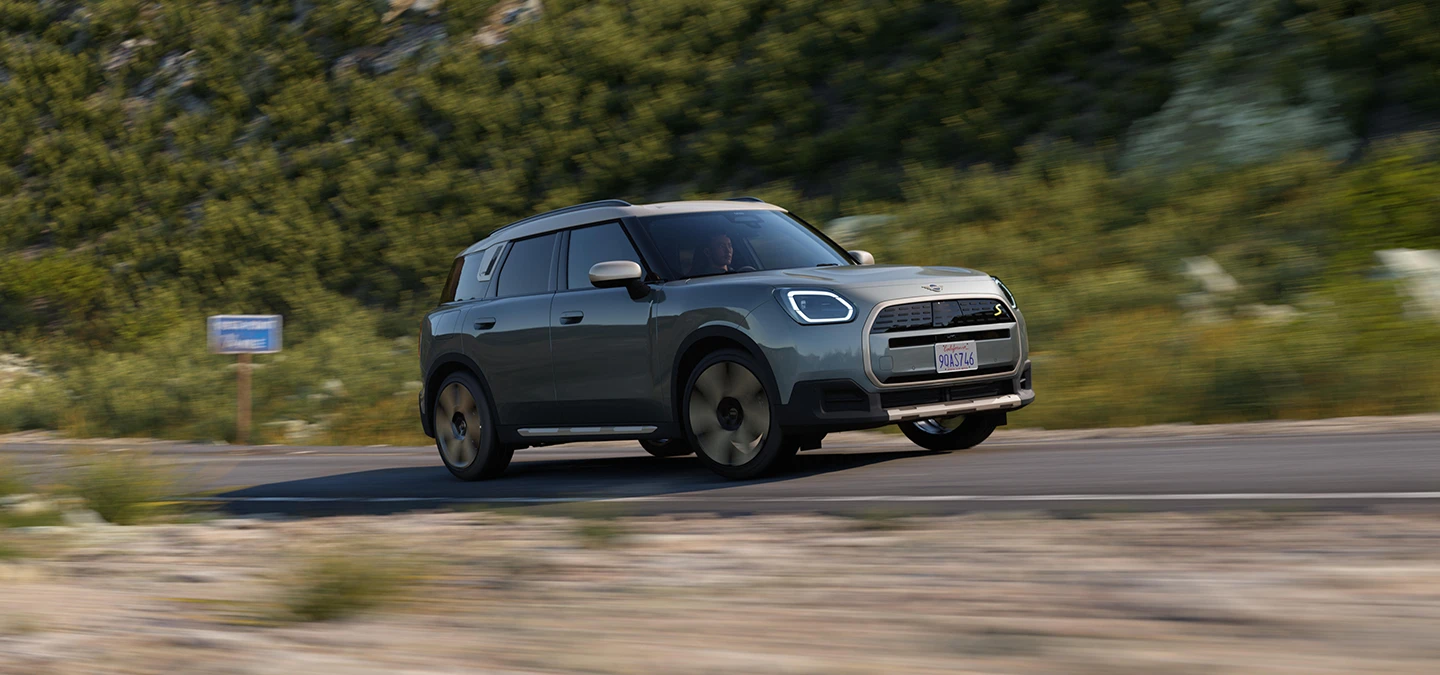 Sānskats uz pilnībā elektrisko MINI Countryman Smokey Green krāsā ar Vibrant Silver jumtu, braucot bezceļa apstākļos.