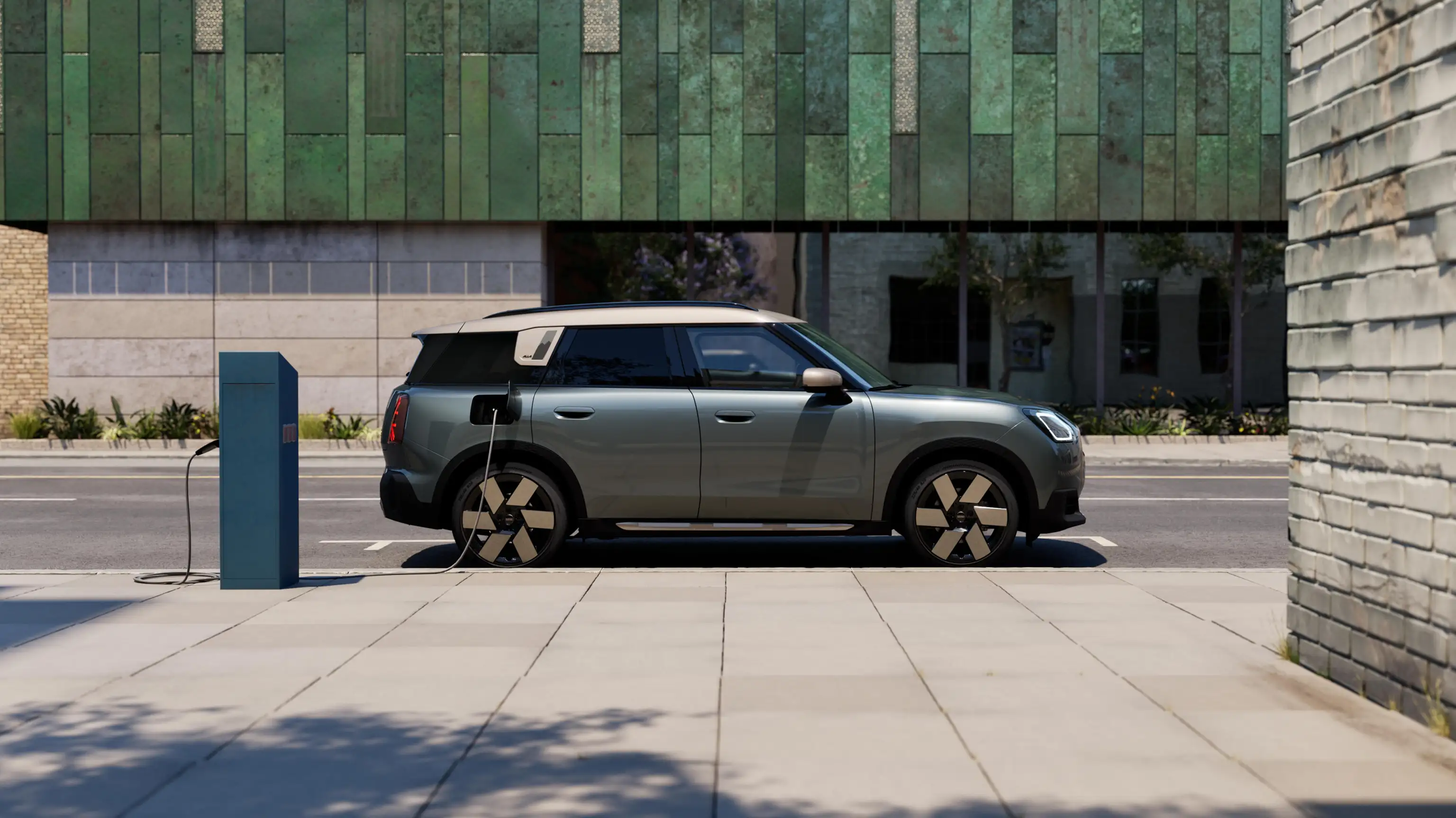 Sānskats – uzlādējošs pilnībā elektriskais MINI Countryman Smokey Green krāsā ar Vibrant Silver jumtu uz ielas