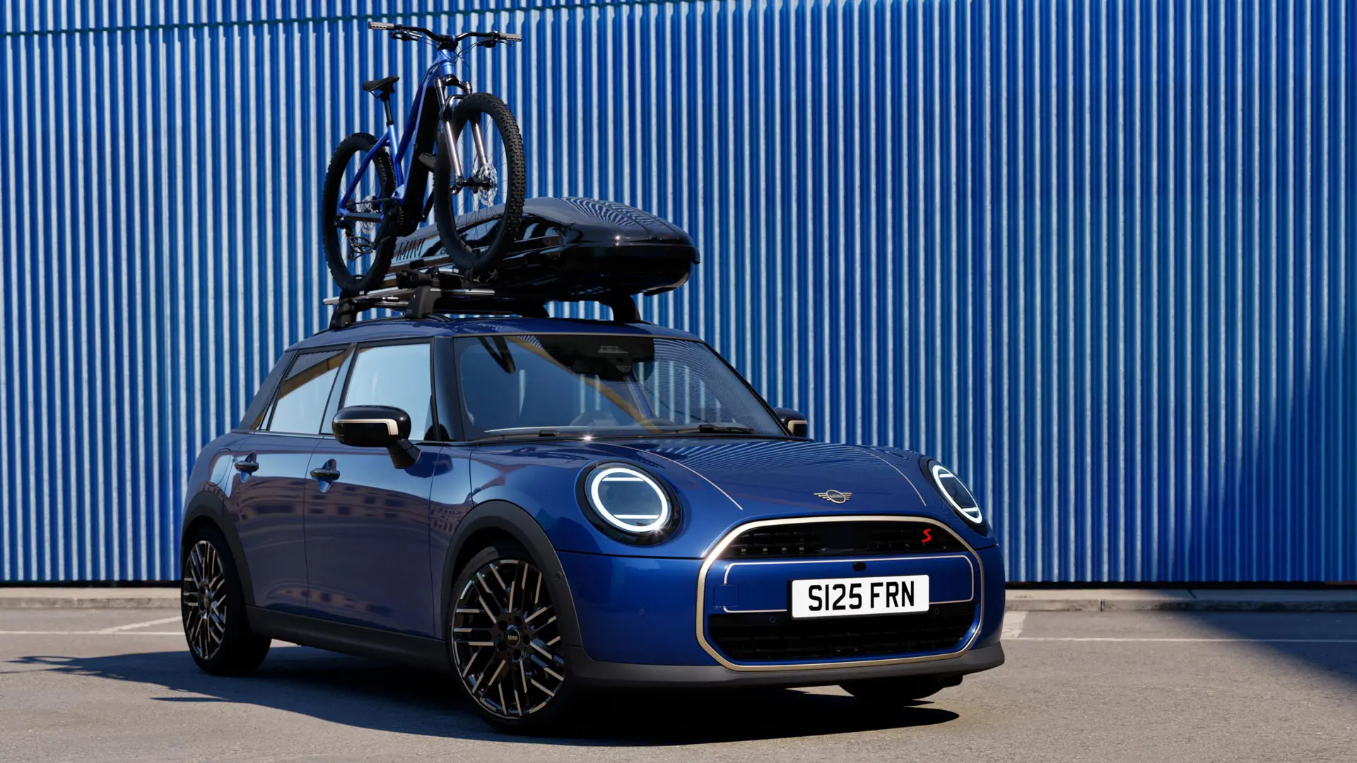 Priekšpuses skats uz MINI Aceman Indigo Sunset Blue krāsā ar MINI jumta velosipēdu bagāžnieku, uz kura atrodas kalnu velosipēds un MINI jumta kaste 320.