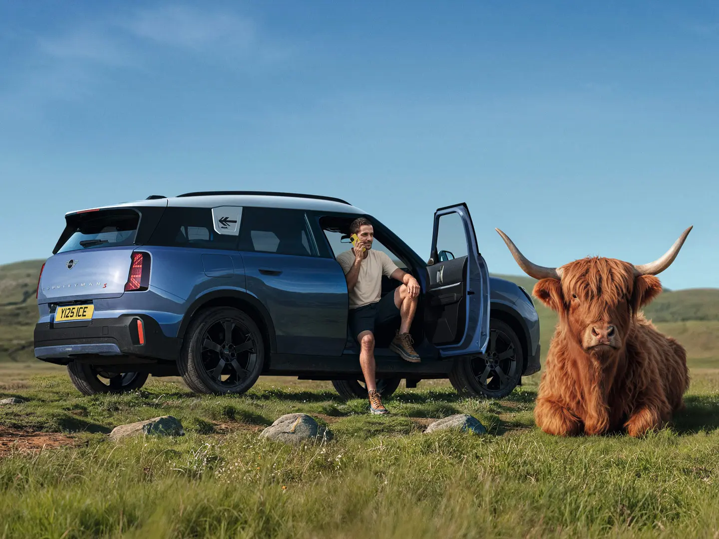 Vīrietis pie sava MINI Countryman S sazinās ar MINI Roadside Assistance, izmantojot MINI lietotni, kamēr netālu guļ Highland bullis.