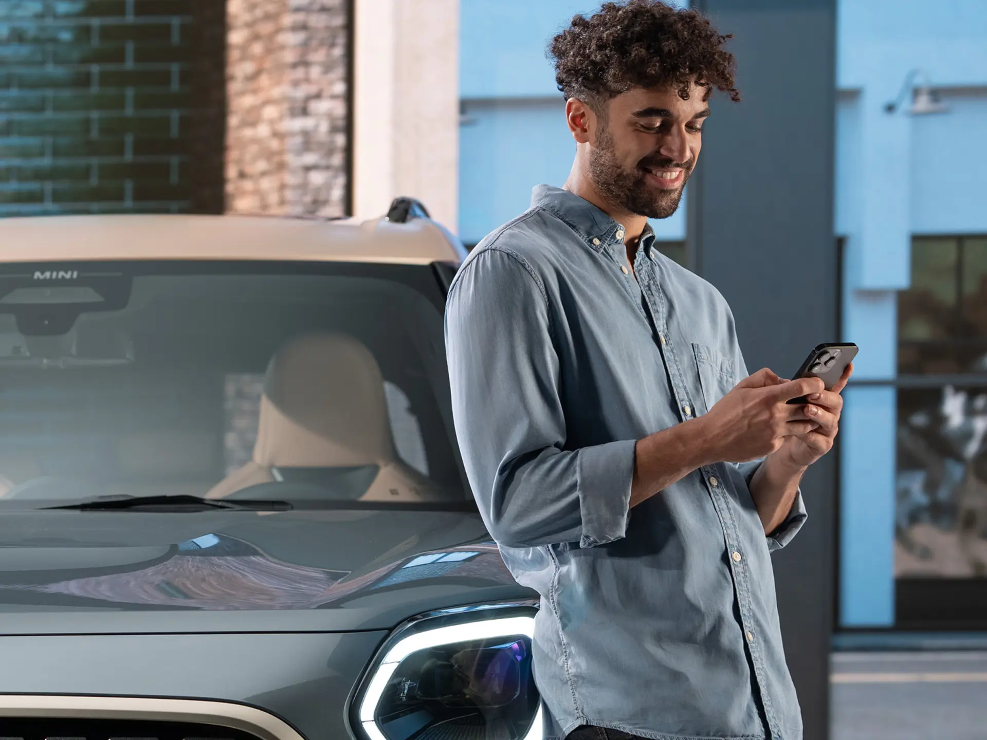 MINI personalizētie servisa pieraksti – vīrietis, kurš blakus MINI automobilim raksta telefonā.