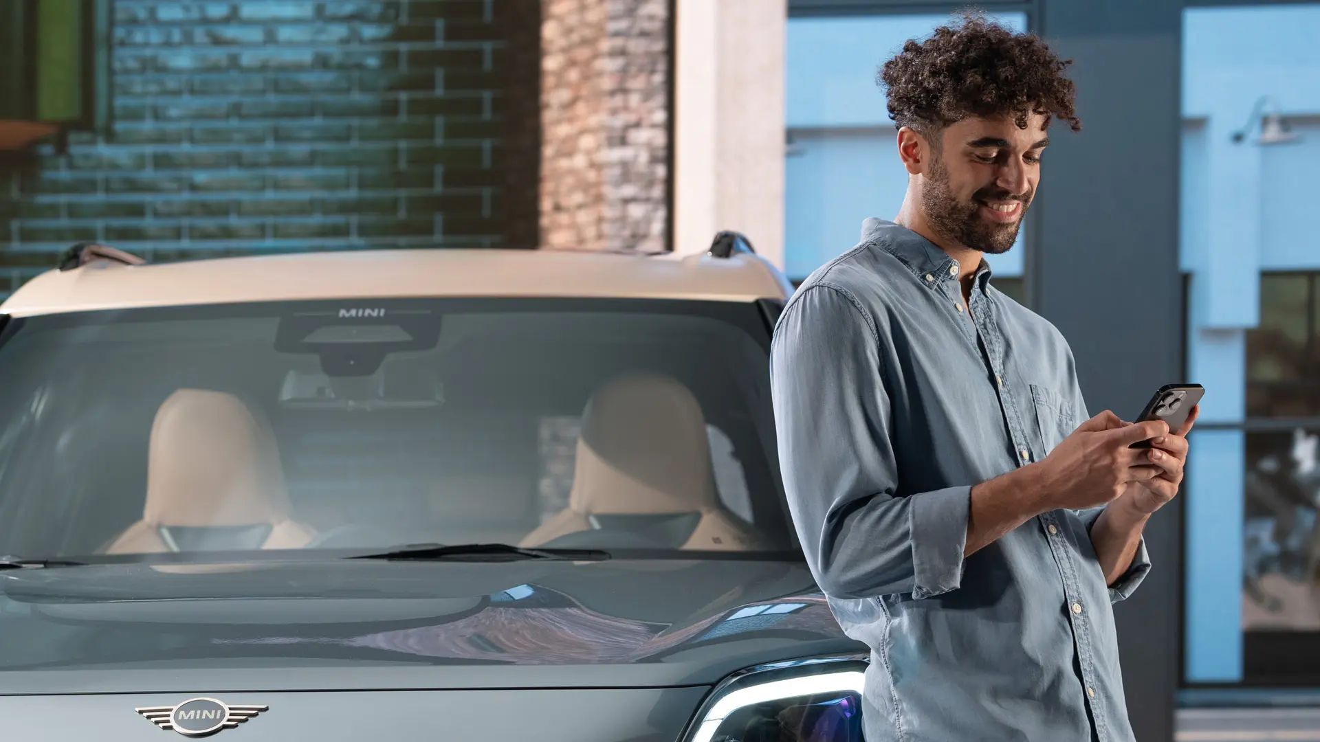 MINI personalizētie servisa pieraksti – vīrietis, kurš blakus MINI automobilim raksta telefonā.